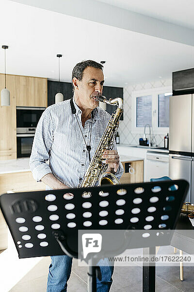 Musician blowing saxophone standing in front of musical stand at home
