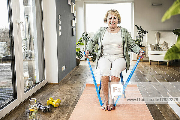 Smiling senior woman with resistance band sitting on chair working out at home