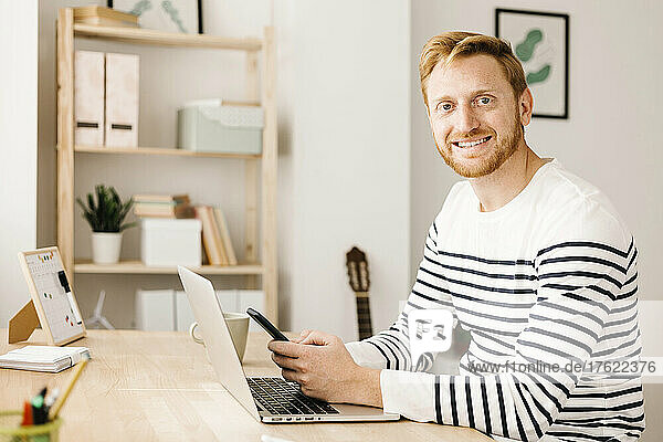 Happy young man holding mobile phone sitting with laptop at table in living room