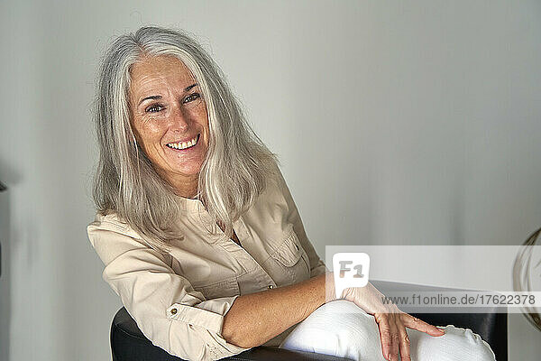 Happy woman with gray hair sitting on chair in front of white wall