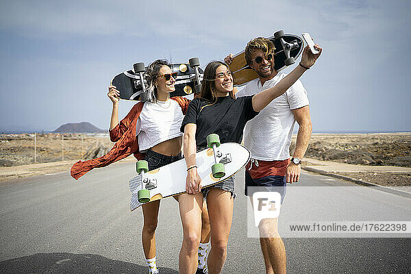 Happy young woman taking selfie with friends holding skateboards through mobile phone on sunny day