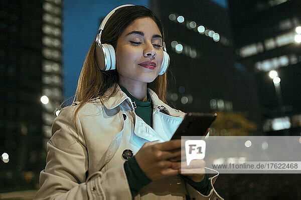 Beautiful woman with eyes closed listening music through wireless headphones at night
