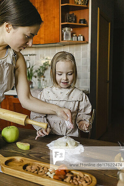 Blond girl looking at mother sprinkling flour on dough in kitchen