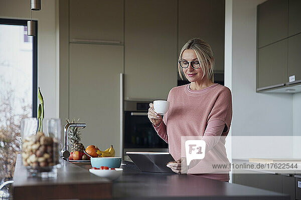 Blond woman holding coffee cup using tablet PC in kitchen