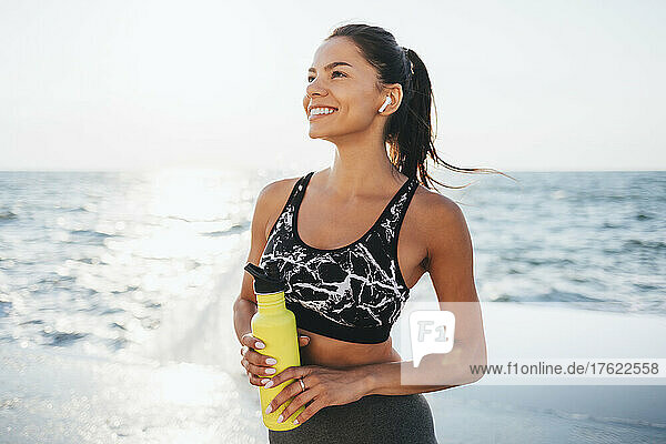 Glückliche junge Frau mit Wasserflasche an einem sonnigen Tag am Strand