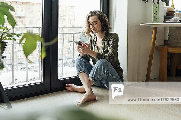 Young woman surfing net through mobile phone by glass window at home