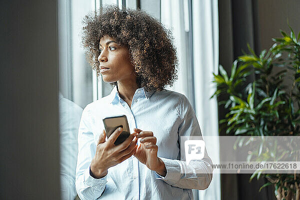 Geschäftsfrau schaut mit Smartphone durchs Fenster im Büro