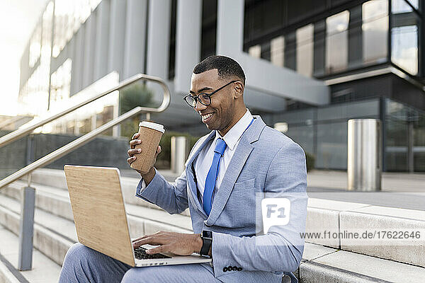 Smiling businessman holding disposable coffee cup using laptop sitting on steps