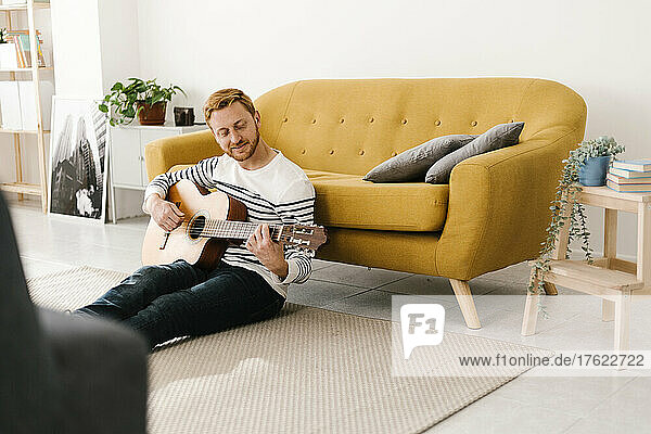 Junger Mann mit roten Haaren übt Gitarre und sitzt zu Hause vor dem Sofa