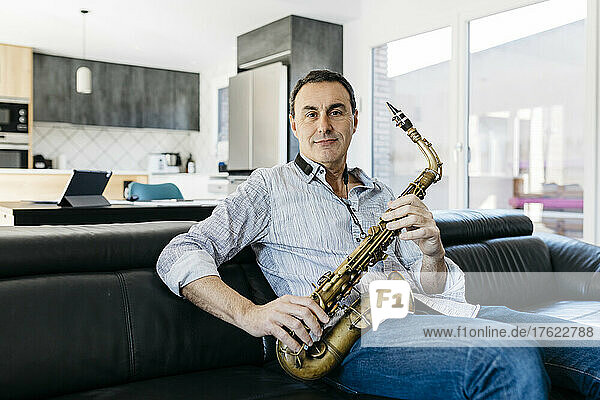 Smiling musician with saxophone sitting on sofa in living room at home