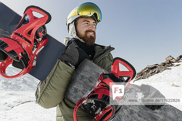 Smiling young man wearing ski goggles holding snowboard on sunny day