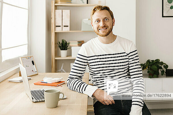 Smiling man sitting with laptop and coffee cup at table in living room