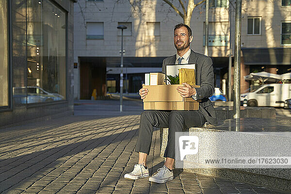 Smiling businessman with box sitting on bench at footpath