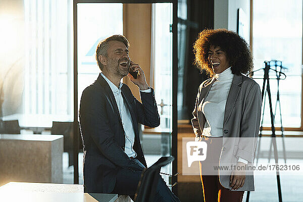 Businessman laughing and talking on mobile phone by colleague at work place