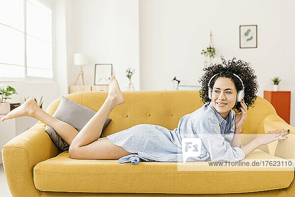 Smiling young woman wearing wireless headphones lying on sofa at home