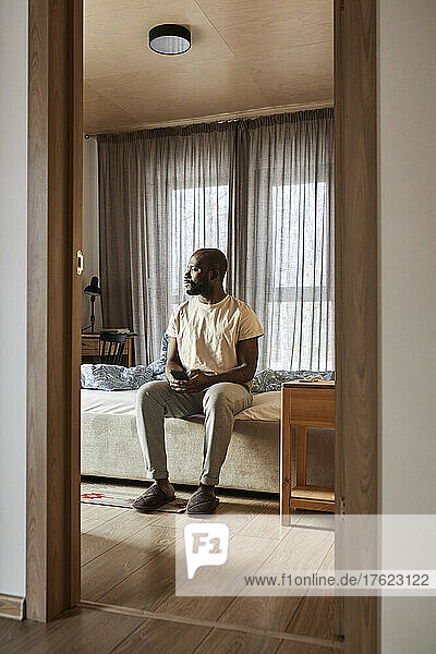 Young man sitting on bed at home seen through doorway