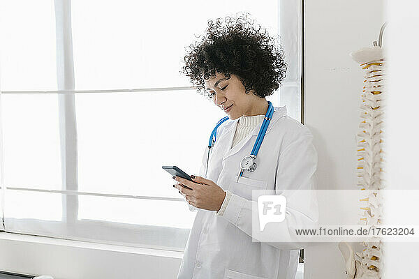 Doctor text messaging through mobile phone standing by window in medical office
