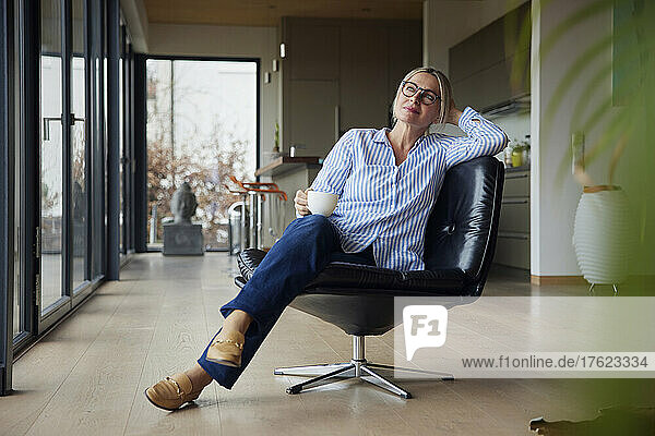 Entspannte Frau mit Brille und Kaffeetasse sitzt zu Hause auf einem Stuhl