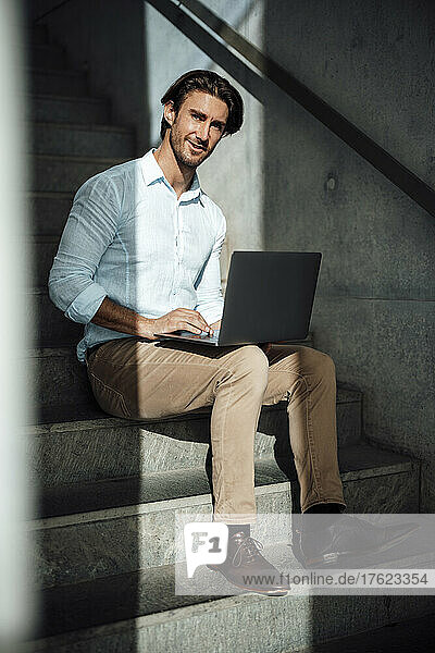 Smiling handsome businessman sitting with laptop on steps