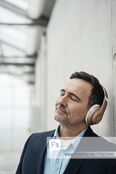 Businessman with eyes closed listening music through wireless headphones by wall