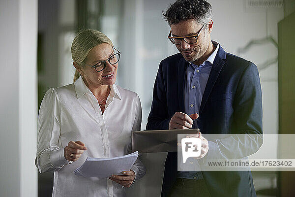 Smiling businesswoman holding document discussing with colleague using tablet PC in office
