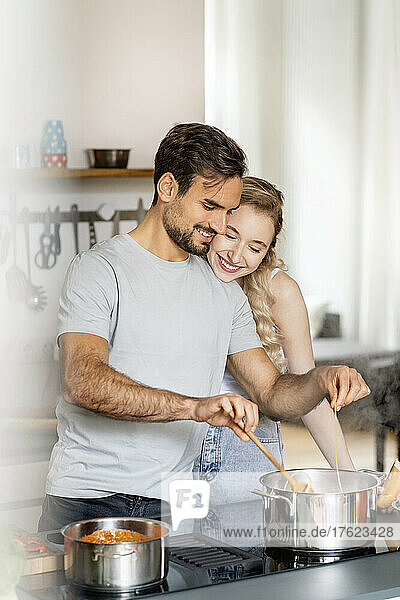 Smiling blond woman embracing boyfriend making pasta in kitchen