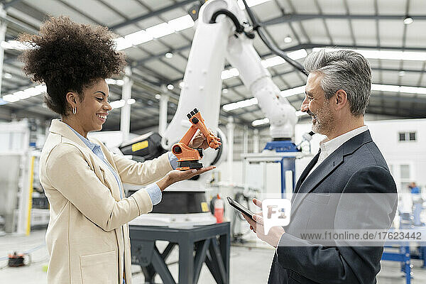 Smiling developer showing model of robotic arm to businessman in factory