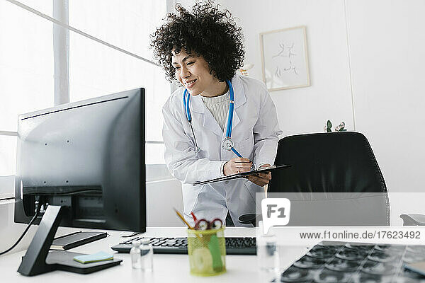 Smiling doctor holding clipboard looking at desktop PC in medical clinic