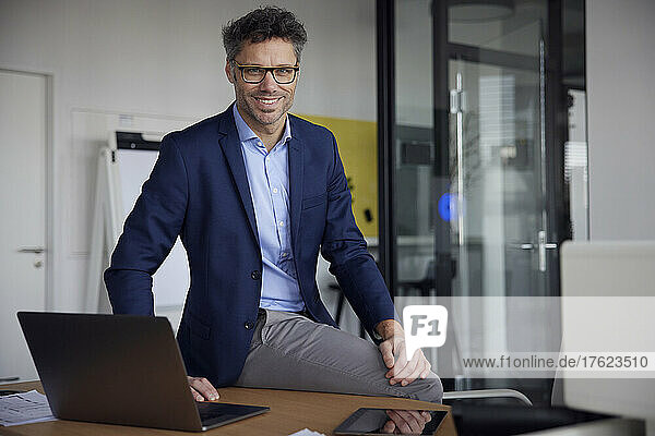 Happy businessman wearing eyeglasses sitting with laptop on desk in office