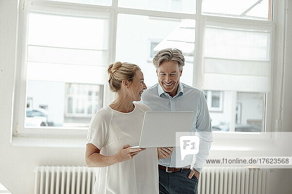 Cheerful businesswoman holding laptop discussing with colleague in office