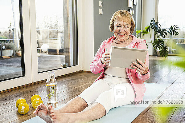 Happy blond senior woman looking at tablet PC on exercise mat at home