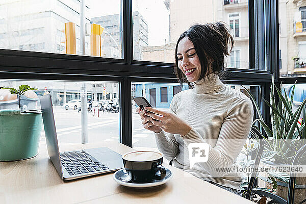 Happy businesswoman with laptop using smart phone sitting in cafe