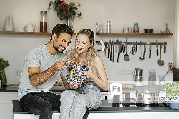 Lächelnde junge Frau blickt auf einen Mann  der zu Hause auf der Kücheninsel Spaghetti isst