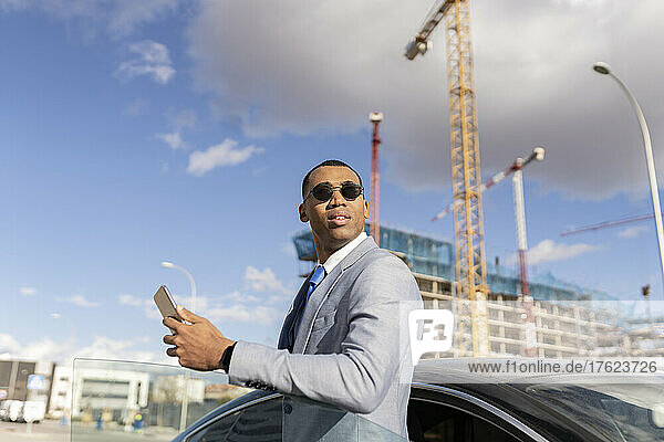 Businessman wearing sunglasses holding smart phone standing by car on sunny day