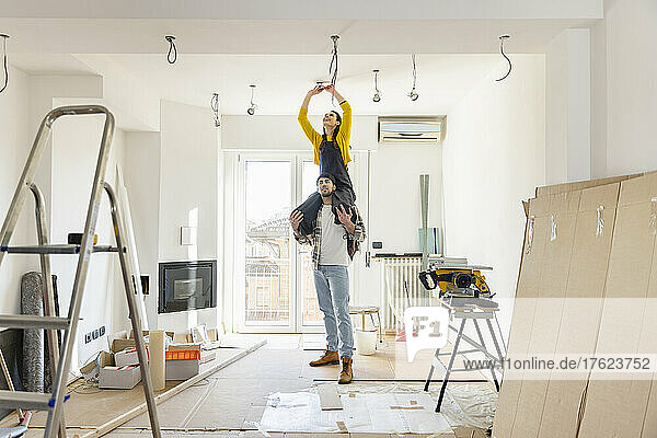 Young man carrying girlfriend on shoulders installing light bulb in living room