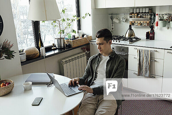 Young freelancer using laptop at table in kitchen