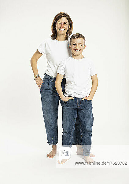 Smiling mother and son standing with hands in pockets against white background