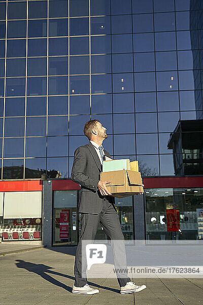 Businessman walking with box of belongings by office building on sunny day