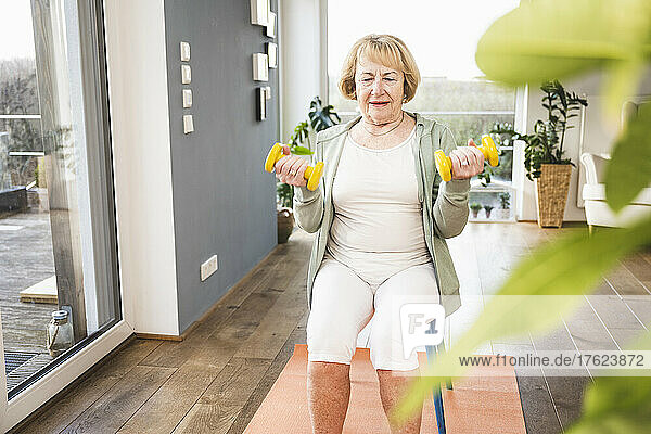 Senior woman with dumbbell sitting on chair exercising at home