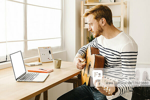Man learning guitar through online tutorial on laptop sitting at table