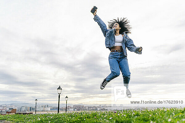 Cheerful woman with smart phone jumping on meadow