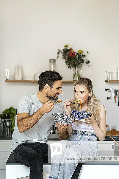 Lächelnder junger Mann schaut seiner Freundin zu  die zu Hause auf der Kücheninsel Spaghetti isst