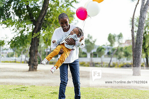 Verspielte Mutter trägt Tochter mit Luftballons im Park
