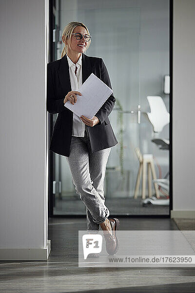 Smiling businesswoman with documents leaning on wall in office