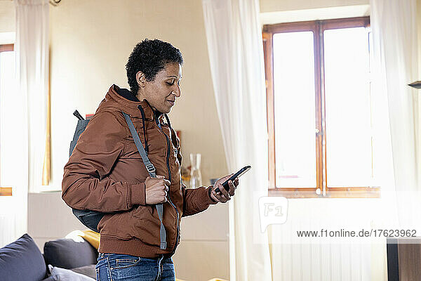 Frau mit Rucksack benutzt Mobiltelefon im heimischen Wohnzimmer