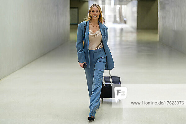 Happy blond businesswoman with wheeled luggage walking at subway station
