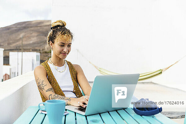 Smiling young woman using laptop sitting at table on rooftop