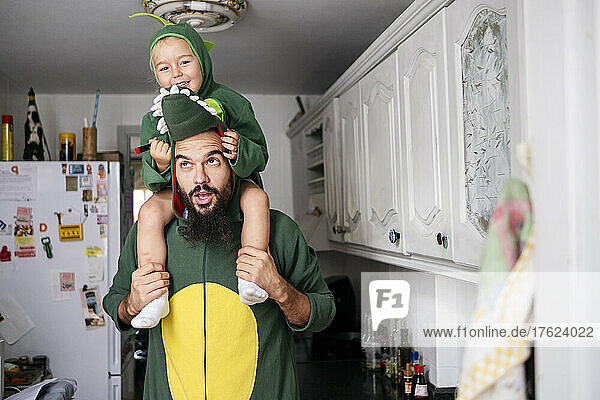 Father carrying boy on shoulders with dinosaur costume in kitchen at home