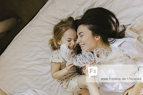 Cheerful girl holding feather lying with mother on bed at home