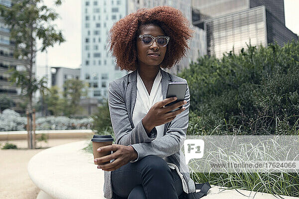 Young businesswoman sitting outdoors with smart phone and disposable coffee cup in hands
